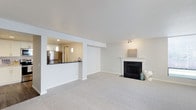 This single-floor residence offers a clean, modern layout designed for comfortable everyday living. The main living area is spacious and filled with natural light, featuring neutral-toned carpeting that adds warmth underfoot and a classic white-mantled fireplace with a sleek black surround as the room's focal point. Recessed and track lighting complement the bright, airy atmosphere created by the white walls and smooth ceilings. The living area flows seamlessly into the kitchen through a built-in pass-through window, which enhances the open feel of the space while maintaining a defined separation between the two areas. The kitchen is well-appointed with dark countertops that contrast cleanly against lighter cabinetry, stainless steel appliances including a refrigerator, and stylish pendant lighting that adds a contemporary touch. The overall ambiance throughout the space is light, neutral, and move-in ready, with a cohesive design palette of whites and soft grays that creates a calm and inviting environment. The thoughtful layout and functional features make this a practical and appealing single-level living space.
