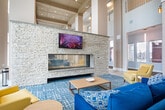Located at 1700 CR-50 in Bay Shore, NY, this impressive community common space at Shoregate is designed to make a grand first impression. The single-floor layout encompasses a generously sized open area that flows seamlessly between a welcoming lounge, hallways, and a sophisticated social space.

The architecture is defined by dramatically soaring double-height ceilings supported by bold structural columns, creating an airy, open atmosphere throughout. Expansive floor-to-ceiling windows line the walls, flooding the interior with abundant natural light and offering clear views of the surrounding community grounds.

A striking stacked-stone fireplace serves as a commanding focal point in the main lounge area, complemented by a wall-mounted flat-screen television above. Comfortable seating — including deep blue sofas and mustard-yellow accent chairs — is arranged atop a patterned teal area rug over light-toned tile flooring, creating an inviting and stylish gathering space.

An adjacent social area features a grand piano alongside dining tables and chairs, underscoring the space's versatility for both casual relaxation and community events. Warm wall sconces and recessed lighting add to the refined ambiance. The overall design palette of soft neutrals, crisp whites, and coastal-inspired blues reflects a modern yet welcoming aesthetic suited to the Bay Shore waterfront community.
