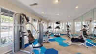 This single-level fitness center is thoughtfully designed to support a comprehensive workout experience. The space is organized into distinct functional zones, accommodating both cardio and strength training activities. Cardio equipment includes multiple treadmills, elliptical trainers, a stationary spin bike, and a rowing machine, while the strength training area features a multi-station cable machine and a cable-and-pulley weight system. A floor-to-ceiling mirror wall runs along one side of the room, effectively doubling the perceived size of the space and allowing users to monitor their form during exercise.

The flooring is a distinctive combination of neutral carpet tiles accented by bold hexagonal patches in varying shades of blue and teal, adding visual energy to the environment. Large, multi-pane windows line the perimeter walls, flooding the interior with natural light and offering views of the surrounding outdoor greenery. Recessed ceiling lighting supplements natural illumination throughout the day. A wall-mounted television is positioned near the cardio section for entertainment during workouts. Ceiling fans and posted fitness center policy signage contribute to the practical, user-focused design of the space.