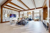 Located at 2926 Barker Cypress Rd in Houston, TX, this sophisticated single-floor community clubhouse and leasing center offers a thoughtfully designed open-concept layout that makes excellent use of its generous footprint. The space is anchored by soaring ceilings adorned with striking exposed wood beams, lending warmth and architectural character to the otherwise bright, contemporary interior. Light-toned herringbone-patterned flooring runs throughout, creating a seamless and polished foundation underfoot. A prominent feature wall clad in layered tile panels serves as a focal backdrop, framing a mounted flat-screen television flanked by round decorative mirrors. Woven rattan pendant chandeliers hang gracefully overhead, casting a warm ambient glow that complements the abundant natural light streaming through floor-to-ceiling windows overlooking lush outdoor greenery and a pool area. The lounge zone is furnished with a rich navy blue sofa, cream accent chairs, and organic wood coffee tables, creating an inviting and well-curated social environment. A dedicated leasing and reception desk station, outfitted with multiple workstations and blue upholstered seating, is seamlessly integrated into the space. Additional areas include a mailroom alcove, a fireplace feature wall, and a corridor leading to further amenities, making this a fully functional and visually impressive community hub.