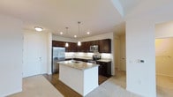 This single-floor space showcases a well-appointed kitchen as its centerpiece. The kitchen features rich, dark espresso-toned cabinetry that extends to the upper walls, providing generous storage throughout. A large central island with a crisp white base is topped with light granite countertops, offering ample prep space and a double undermount sink with a modern arc faucet.

The granite countertops continue along the perimeter of the kitchen, complementing the full suite of stainless steel appliances, which includes a French door refrigerator with an ice and water dispenser, an electric range, and an over-the-range microwave. Recessed lighting is complemented by two elegant pendant lights that hang above the island, creating focused task lighting.

The kitchen transitions seamlessly into an adjacent carpeted living area, suggesting an open-concept layout. Hardwood flooring in the kitchen contrasts with the plush carpeting of the surrounding rooms, clearly defining the kitchen space. The neutral walls and clean white ceilings contribute to a bright, modern aesthetic throughout the space.