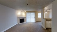 This single-floor living space presents a comfortable and well-appointed interior designed for everyday living. The main living area is generously sized, anchored by a classic fireplace with a clean white surround and black firebox, serving as a natural focal point within the room. A textured popcorn ceiling adds dimension overhead, while track lighting provides adjustable, directed illumination along one wall. Plush wall-to-wall carpeting in a neutral tone runs throughout the space, contributing to a warm and inviting atmosphere. A full-height sliding glass door dressed with vertical blinds allows natural light to filter in and provides access to an outdoor area, while an additional window on the adjacent wall further brightens the interior. The warm beige wall tones complement the neutral flooring and create a cohesive, understated palette throughout. A partial wall or bar-height partition is visible toward one side of the space, suggesting a transition into a kitchen or dining area. The overall layout is open and functional, well-suited for a variety of furniture arrangements.