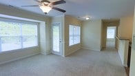 This single-floor space offers a welcoming and well-lit interior designed for comfortable living. The main living area is finished with plush wall-to-wall carpeting that adds warmth underfoot, while soft yellow-toned walls create a calm and neutral backdrop throughout. Crown molding runs along the upper edges of the walls, lending a classic, refined touch to the space.

Natural light is a defining feature, with multiple windows dressed in white horizontal blinds positioned throughout the room. A glass-paned exterior door provides both an additional source of daylight and a direct connection to the outdoors, framed by views of surrounding greenery. A ceiling fan with an integrated light fixture is centrally mounted overhead, ensuring both air circulation and ambient lighting.

Chair rail molding adds a subtle architectural detail along the lower portion of the walls, contributing to the traditional character of the space. Recessed lighting supplements the natural light in areas further from the windows, maintaining a bright and even illumination throughout. Clean white baseboards and trim provide a crisp contrast to the warm wall tones, completing the polished, move-in-ready feel of this thoughtfully detailed space.