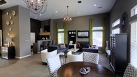 This single-story space is designed as a sophisticated communal lounge or clubroom, offering a well-appointed interior that balances modern aesthetics with comfortable functionality. The open-concept layout accommodates several distinct seating arrangements, including dark upholstered sofas accented with decorative pillows, curved barrel chairs upholstered in chevron-patterned fabric, and a dark wood dining table, creating versatile zones for relaxing or socializing.

Two globe-style chandelier light fixtures with exposed bulbs serve as eye-catching focal points, complementing the recessed lighting throughout the ceiling. The walls are finished in a deep charcoal tone, providing a dramatic backdrop that contrasts with the vibrant lime-green drapery panels framing multiple large windows. These windows allow generous natural light to fill the space while offering views of the surrounding greenery.

A kitchenette area toward the rear includes a bar-height counter with seating, cabinetry, and countertop appliances, making the space suitable for gatherings and events. A wall-mounted flat-screen television adds a functional entertainment element. The overall ambiance is polished, contemporary, and inviting.