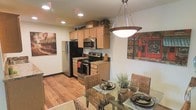 This single-story space seamlessly integrates the kitchen and dining area into a cohesive, open-concept layout. The kitchen is well-appointed with stainless steel appliances, including a refrigerator, electric range, and built-in microwave. Light wood cabinetry extends to the upper walls, offering generous storage, while dark granite countertops provide a striking contrast and durable workspace. Recessed lighting illuminates the kitchen, enhancing the warm tones of the hardwood flooring that runs throughout the space.

The dining area transitions naturally from the kitchen, anchored by a glass-top table surrounded by woven rattan chairs that add a relaxed, organic texture to the room. A bold pendant light fixture hangs above the dining table, serving as a focal point and providing targeted illumination for meals. Decorative wall art featuring urban café scenes adds a lively, cosmopolitan character to the space. Potted greenery on the dining table introduces a natural element, complementing the overall warm and welcoming ambiance of this thoughtfully designed living space.