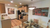 This single-story space seamlessly integrates the kitchen and dining area into a cohesive, open-concept layout. The kitchen is well-appointed with stainless steel appliances, including a refrigerator, electric range, and built-in microwave. Light wood cabinetry extends to the upper walls, offering generous storage, while dark granite countertops provide a striking contrast and durable workspace. Recessed lighting illuminates the kitchen, enhancing the warm tones of the hardwood flooring that runs throughout the space.

The dining area transitions naturally from the kitchen, anchored by a glass-top table surrounded by woven rattan chairs that add a relaxed, organic texture to the room. A bold pendant light fixture hangs above the dining table, serving as a focal point and providing targeted illumination for meals. Decorative wall art featuring urban café scenes adds a lively, cosmopolitan character to the space. Potted greenery on the dining table introduces a natural element, complementing the overall warm and welcoming ambiance of this thoughtfully designed living space.
