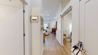 This well-appointed single-floor residence in Medfield, Massachusetts offers a thoughtfully designed layout that maximizes comfort and functionality. The open kitchen is a standout feature, showcasing two-tone cabinetry — warm wood lower cabinets paired with crisp white uppers — accented by gold hardware, a gray subway tile backsplash, white quartz countertops, stainless steel appliances, and a generous island with seating for casual dining. Pendant lighting adds a refined touch overhead. The combined living and dining area is bathed in natural light from a row of windows dressed with sheer café curtains, and features light wood-look flooring that flows throughout. The living space offers a warm, cottage-inspired ambiance with a classic sofa, framed botanical artwork, and a charming dining nook. The bedroom is a cozy retreat with clean white walls, a classic metal-frame bed, and ample storage. The bathroom is elegantly finished with floor-to-ceiling marble-look tile, a frameless glass sliding shower enclosure, and clean white surfaces that lend a spa-like feel. This residence blends modern finishes with timeless, livable charm.