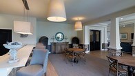 This single-floor space presents a refined and welcoming interior that balances classic design with comfortable functionality. The dining area serves as a central focal point, anchored by a round wooden table surrounded by cross-back chairs, all set atop a neutral area rug that adds warmth and definition to the space. A rustic wooden sideboard lines the wall, styled with decorative candelabras and arched wicker accents, while a teal mosaic-framed circular mirror above it introduces a subtle pop of color against the soft sage-toned walls.

Overhead, large drum pendant lights hang at staggered heights, casting a warm, even glow throughout the room and contributing to the space's inviting ambiance. To one side, a row of upholstered chairs with nailhead trim detail suggests an additional lounge or seating area, ideal for social gatherings. Stone veneer accents near the entryway add a natural, grounded texture that complements the wood and neutral tones found throughout.

The flooring transitions between hardwood and area rugs, guiding movement through the space naturally. Dark-framed double doors at the rear provide a strong architectural contrast and lead to adjacent areas, reinforcing the space's open, connected layout. Overall, the design exudes a casual elegance suited for both relaxed and social settings.