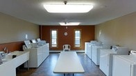 This shared laundry facility is designed for practicality and efficiency. The room is finished with a warm, deep brown accent wall that contrasts with the lighter surrounding walls, creating a functional yet visually defined space. Two sets of washers and dryers line opposite walls — one set on the left and another on the right — providing ample capacity for multiple users simultaneously. A long folding table runs down the center of the room, offering a convenient surface for sorting and folding laundry. The tile flooring is durable and easy to maintain, well-suited to the demands of a high-use laundry environment. Overhead, a large flush-mounted fluorescent light panel and a ceiling fan with an additional skylight panel ensure the room is well-lit and ventilated. Two wood-trimmed windows on the back wall allow natural light to filter into the space and provide views of the exterior. A wall-mounted clock keeps track of laundry cycles, and a small chair is available for those waiting. Baseboard heating along the rear wall helps maintain comfort during colder months.