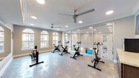 This fitness studio is thoughtfully designed to support a variety of cardio-focused workouts in a bright, polished environment. The space is organized into distinct functional zones, beginning with a cycling and rowing area outfitted with multiple stationary bikes and a rowing machine. Arched windows along one wall flood this section with natural light while offering views of the surrounding exterior, creating an energizing atmosphere during workouts.

A full wall of floor-to-ceiling mirrors runs along one side of the studio, visually expanding the space and allowing users to monitor their form. Ceiling fans are positioned throughout to maintain airflow and comfort, complemented by recessed lighting that ensures consistent illumination. Crown molding and clean white trim lend the studio a refined, upscale character that sets it apart from a standard gym environment.

Adjacent to the main exercise area, a support zone features a wall-mounted flat-screen television, a compact under-counter refrigerator, and dark cabinetry with a light countertop — providing a convenient station for hydration and rest between sessions. A glass-paneled door connects this area to an adjoining space, hinting at additional amenities nearby. The durable, patterned flooring throughout is designed to withstand heavy equipment use while maintaining an attractive finish.