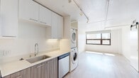 This thoughtfully laid-out 1-bedroom, 1-bathroom unit offers a modern, efficient design across a single floor. The open-concept kitchen is a standout feature, showcasing two-tone cabinetry with white upper cabinets and wood-grain lower cabinets, stainless steel appliances including a range, over-the-range microwave, dishwasher, and full-size refrigerator, all complemented by clean white countertops and under-cabinet lighting. A stacked washer and dryer unit is conveniently integrated into the kitchen area, adding practical in-unit laundry access. Light wood-style flooring runs throughout the main living areas, lending warmth and continuity to the space. The living area benefits from a large picture window that invites generous natural light and offers sweeping urban city views. The bedroom is a calm, minimalist retreat featuring the same light hardwood flooring, clean white walls, and a wide triple-panel sliding closet that maximizes storage. The bathroom is finished to a high standard with a wood-toned floating vanity, white quartz countertop, framed mirror, stylish wall-mounted lighting, and a glass-enclosed walk-in shower with white subway tile. Industrial-style track lighting on the ceiling adds a contemporary edge throughout the unit, enhancing the overall modern urban ambiance.
