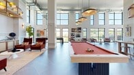 Located in Brentwood, Tennessee, this thoughtfully designed community amenity space offers a sophisticated blend of social and recreational areas across a generously sized single floor. Soaring double-height ceilings define the main gathering area, which is anchored by a statement billiards table illuminated by a cluster of gold pendant lights. Large black-framed windows line the walls, flooding the interior with natural light and framing panoramic views of the surrounding Tennessee hillside landscape.

The lounge area features plush rust-colored armchairs and a tufted green banquette booth, creating intimate seating vignettes accented by curated gallery wall arrangements. A dedicated dining zone provides casual bar-height seating alongside a high-top table, while a fully equipped kitchen area is outfitted with dark cabinetry, marble countertops with brass-legged island, a tiled backsplash, and direct access to an outdoor balcony overlooking the pool area.

A mezzanine level with warm wood accents and black iron railings overlooks the main floor, adding vertical drama and architectural depth. Large-format slate-toned tile flooring laid in a herringbone pattern ties the space together, complementing the modern-industrial aesthetic. A reception desk near the entrance completes this well-appointed community hub.