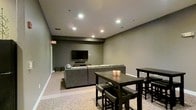 This single-floor space is designed as a comfortable communal lounge and entertainment area. The room features neutral gray walls and smooth ceilings fitted with recessed pot lights, which cast a warm, even glow throughout the space. The flooring transitions between hard surface and carpet, subtly delineating the different functional zones within the room.

The media area anchors one end of the space, centered around a wall-mounted flat-screen television paired with a sound bar. A generously sized gray sectional sofa faces the screen, offering ample seating for gatherings or casual viewing. The arrangement creates an inviting, relaxed atmosphere suited for social use.

Adjacent to the lounge area, two dark-toned dining tables with matching chairs and stools provide additional seating and surface space, extending the room's versatility for dining or group activities. Candle holders on one of the tables add a subtle decorative touch.

An emergency exit sign is visible near the entrance, indicating this is a shared or amenity-style space. Overall, the room presents a functional, modern aesthetic with a muted, understated color palette that promotes a calm and social environment.
