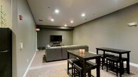This single-floor space is designed as a comfortable communal lounge and entertainment area. The room features neutral gray walls and smooth ceilings fitted with recessed pot lights, which cast a warm, even glow throughout the space. The flooring transitions between hard surface and carpet, subtly delineating the different functional zones within the room.

The media area anchors one end of the space, centered around a wall-mounted flat-screen television paired with a sound bar. A generously sized gray sectional sofa faces the screen, offering ample seating for gatherings or casual viewing. The arrangement creates an inviting, relaxed atmosphere suited for social use.

Adjacent to the lounge area, two dark-toned dining tables with matching chairs and stools provide additional seating and surface space, extending the room's versatility for dining or group activities. Candle holders on one of the tables add a subtle decorative touch.

An emergency exit sign is visible near the entrance, indicating this is a shared or amenity-style space. Overall, the room presents a functional, modern aesthetic with a muted, understated color palette that promotes a calm and social environment.