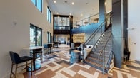 This community clubhouse is defined by its dramatic architectural presence, anchored by soaring double-height ceilings that flood the main gathering area with natural light through expansive floor-to-ceiling windows. A bold geometric-patterned tile floor in neutral tones of tan, gray, and white sets a sophisticated foundation throughout the open-plan space, immediately establishing a contemporary design identity.

A steel-cable staircase with an industrial-modern aesthetic leads to an upper mezzanine level, where bar-height seating and additional gathering space overlook the main floor below. Pendant globe lighting cascades from above, complementing recessed lighting throughout. A billiards table occupies its own distinct zone, adding a recreational focal point near the staircase.

The lounge area transitions seamlessly into a more intimate entertainment zone, featuring a large wall-mounted television framed by a striking diamond-patterned tile accent wall. Plush sectional sofas, armchairs, and layered rugs in a coordinated blue, gray, and taupe palette create an inviting, well-curated atmosphere. Smaller seating alcoves offer quieter spaces for individual use. Large glass doors throughout connect the interior to the surrounding outdoor environment, enhancing the sense of openness.