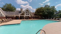 Located at 4300 Furman Hall Rd in Raleigh, NC, the Highlands at Raleigh amenity area is a thoughtfully designed outdoor community space that offers a range of recreational and social features. At its centerpiece is a generously sized, freeform swimming pool with clear turquoise water, surrounded by a spacious concrete pool deck lined with lounge chairs for sunbathing. The pool is flanked by a covered brick patio supported by substantial dark brick columns, providing shaded seating and a welcoming transition between the indoors and the pool area. Classic white rocking chairs positioned along the covered porch offer a charming, relaxed gathering spot. The clubhouse building, featuring a steeply pitched roofline, prominent chimney, and large multi-pane windows, serves as an architectural anchor to the space. A separate outdoor lounge area is appointed with comfortable cushioned seating arranged around a modern fire pit table, set within a landscaped garden enclosed by iron fencing and mature trees. An additional pergola-covered grilling station, constructed with brick and granite countertops and equipped with stainless steel grills, provides an ideal space for outdoor cooking. Together, these amenities create a vibrant, resort-inspired outdoor environment ideal for relaxation and community gatherings.