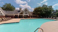 Located at 4300 Furman Hall Rd in Raleigh, NC, the Highlands at Raleigh amenity area is a thoughtfully designed outdoor community space that offers a range of recreational and social features. At its centerpiece is a generously sized, freeform swimming pool with clear turquoise water, surrounded by a spacious concrete pool deck lined with lounge chairs for sunbathing. The pool is flanked by a covered brick patio supported by substantial dark brick columns, providing shaded seating and a welcoming transition between the indoors and the pool area. Classic white rocking chairs positioned along the covered porch offer a charming, relaxed gathering spot. The clubhouse building, featuring a steeply pitched roofline, prominent chimney, and large multi-pane windows, serves as an architectural anchor to the space. A separate outdoor lounge area is appointed with comfortable cushioned seating arranged around a modern fire pit table, set within a landscaped garden enclosed by iron fencing and mature trees. An additional pergola-covered grilling station, constructed with brick and granite countertops and equipped with stainless steel grills, provides an ideal space for outdoor cooking. Together, these amenities create a vibrant, resort-inspired outdoor environment ideal for relaxation and community gatherings.