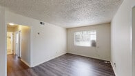 This single-story space offers a straightforward, functional layout across several rooms, as revealed by the 3D floor plan model. The main living area features durable wood-look laminate flooring in a gray-toned finish, crisp white walls, and a textured popcorn ceiling. A window fitted with horizontal blinds allows natural light to enter while providing views of surrounding greenery. A wall-mounted air conditioning unit provides supplemental climate control alongside a ceiling vent.

The bedrooms are finished with neutral wall-to-wall carpeting and feature their own windows with blinds. One bedroom offers two windows positioned on adjacent walls, bringing in generous natural light and views of the exterior brick surroundings, while another looks out onto a grassy outdoor area.

The kitchen is equipped with white cabinetry above and below, dark granite-style laminate countertops, a stainless steel sink, and a white gas range with four burners. A window above the counter area adds brightness to the space.

The bathroom features classic white ceramic tile with a bold navy accent border, a pedestal-style sink with chrome fixtures, a mirrored medicine cabinet, and a tub-shower combination. A small window provides natural ventilation. Throughout, the space maintains a clean, utilitarian character.