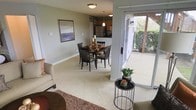 This single-floor residence is designed with an open-concept floor plan that promotes a natural flow between the primary living spaces. The living area features plush wall-to-wall carpeting, neutral gray-toned walls, and is anchored by a cozy seating arrangement accented with layered area rugs. A framed landscape artwork adds a finishing touch to the living space.

The dining area sits centrally within the open layout, accommodating a dark-stained wooden dining table with matching chairs, illuminated by a pendant light overhead. This placement allows for easy interaction between the dining and adjacent kitchen spaces.

The kitchen is positioned at the rear of the open plan and is outfitted with warm wood cabinetry, a stainless steel refrigerator, and overhead pendant lighting that adds a welcoming ambiance.

One of the standout features of this residence is the sliding glass door that opens directly to a covered outdoor patio, offering views of lush greenery beyond. This seamless indoor-outdoor connection allows abundant natural light to filter into the living and dining areas, enhancing the overall brightness and airy feel of the space.