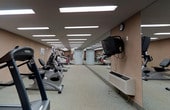 This fitness center offers a dedicated, single-floor space designed to support a comprehensive workout experience. The room features a generous layout that accommodates an array of cardio equipment, including treadmills, ellipticals, and stationary bikes, all arranged in an orderly fashion to maximize usability and flow. Bright recessed fluorescent panel lighting runs the length of the ceiling, ensuring the space is evenly and thoroughly illuminated throughout. A full-length mirror along one wall visually expands the room while allowing users to monitor their form during exercise. A wall-mounted television is positioned conveniently for viewing during workouts, adding to the overall comfort of the space. The flooring consists of a durable, speckled surface well-suited for high-traffic fitness use. Warm-toned walls contribute to an energizing yet functional atmosphere. A secondary door at the far end of the room provides additional access, enhancing circulation through the space. The combination of practical design, ample equipment, and thoughtful amenities makes this fitness center a fully functional and inviting facility for daily exercise.