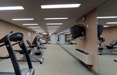 This fitness center offers a dedicated, single-floor space designed to support a comprehensive workout experience. The room features a generous layout that accommodates an array of cardio equipment, including treadmills, ellipticals, and stationary bikes, all arranged in an orderly fashion to maximize usability and flow. Bright recessed fluorescent panel lighting runs the length of the ceiling, ensuring the space is evenly and thoroughly illuminated throughout. A full-length mirror along one wall visually expands the room while allowing users to monitor their form during exercise. A wall-mounted television is positioned conveniently for viewing during workouts, adding to the overall comfort of the space. The flooring consists of a durable, speckled surface well-suited for high-traffic fitness use. Warm-toned walls contribute to an energizing yet functional atmosphere. A secondary door at the far end of the room provides additional access, enhancing circulation through the space. The combination of practical design, ample equipment, and thoughtful amenities makes this fitness center a fully functional and inviting facility for daily exercise.