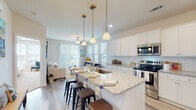 This single-story home is designed around an open-concept layout that seamlessly connects the kitchen, dining, and living areas, creating a cohesive and functional living environment. The kitchen serves as a central focal point, featuring white shaker-style cabinetry, a classic subway tile backsplash, and light granite countertops that extend across a generously sized island with seating for multiple bar stools. Three pendant lights with brass accents hang above the island, complementing the stainless steel appliances, which include a refrigerator, range, and built-in microwave. Light wood-tone flooring flows throughout the main living areas, enhancing the bright, airy atmosphere.

The living room benefits from an abundance of natural light provided by large windows and a sliding glass door, both dressed with white blinds. A ceiling fan is centrally positioned overhead. The space connects to a sunlit sitting area framed by additional windows offering outdoor views.

The bedrooms are carpeted for comfort and each include ceiling fans. The primary bedroom features direct access to a private bathroom equipped with a single-sink vanity, soaking tub, and light wood-tone flooring. A secondary bedroom is equally well-lit with a window and neutral finishes throughout.