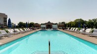 Located at 5252 Riggins Run in Dublin, Ohio, this outdoor amenity space is designed to deliver a resort-like experience for its community. The centerpiece is a generously sized, crystal-clear swimming pool with vivid turquoise water, framed by a broad, well-maintained concrete sundeck. Rows of white lounge chairs line both sides of the pool, offering ample space for sunbathing and relaxation. Striped umbrellas are positioned throughout the deck, providing convenient shade options for guests.

At the far end of the pool, a handsome pavilion serves as the architectural focal point of the space. Built with a classic hip roof and warm stone masonry, the structure houses what appears to be a media or lounge area, making it ideal for social gatherings. Flanking the pavilion are symmetrical pergola-style structures with billowing white curtain panels, creating a series of semi-private cabana retreats that blend elegance with comfort. Lush mature trees form a natural green backdrop, adding privacy and a sense of seclusion to the entire space. The open sky above ensures abundant natural light throughout the day, enhancing the vibrant and inviting atmosphere of this well-appointed outdoor destination.