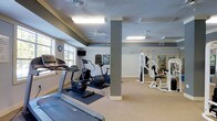 This fitness center offers a functional and well-organized space designed to support a full range of workout routines. The layout is open and spacious, with a neutral color palette of soft grays and whites that creates a clean, focused environment. Carpeted flooring covers the main exercise area, providing a comfortable surface underfoot.

The cardio section is outfitted with a treadmill, elliptical trainer, and stationary bike, each positioned to take advantage of the natural light streaming in through large windows along one wall. A wall-mounted television is conveniently placed nearby for entertainment during workouts. Ceiling fans are installed throughout to promote airflow and comfort during exercise.

On the strength training side, a multi-station cable machine offers a variety of resistance exercises, making it suitable for users at different fitness levels. A structural column divides the space subtly without impeding movement or sightlines between zones. Mirrors along one wall help maximize the sense of space while allowing users to monitor their form.

The facility is well-lit with overhead fluorescent lighting, and safety signage is clearly posted throughout. An adjacent area, visible through a doorway, suggests additional amenity spaces nearby. Overall, the fitness center provides a practical, comfortable, and fully equipped environment for daily exercise.