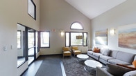 This single-floor space makes a striking first impression with its expansive lobby lounge, defined by soaring vaulted ceilings that lend an open, airy quality to the interior. A signature arched window crowns the wall above a trio of large rectangular windows, flooding the space with natural light and offering views of the surrounding greenery outside. The tall, neutral-toned walls amplify the sense of height and openness throughout.

The lounge area is thoughtfully appointed with contemporary furnishings, including a plush grey sectional sofa accented with warm orange throw pillows, a pair of sleek amber leather accent chairs, and a set of round marble-topped coffee tables with chrome bases. A large-scale abstract artwork adorns one wall, adding a refined artistic touch, while a wall-mounted flat-screen television provides a functional amenity. Stylish pendant and wall-mounted light fixtures complement the natural light and contribute to a welcoming ambiance.

The flooring transitions from smooth tile at the entrance to a patterned area rug beneath the seating arrangement, anchoring the lounge zone and adding textural depth. Overall, the space balances a sophisticated, modern aesthetic with a comfortable and inviting atmosphere, making it well-suited for gathering and relaxation.