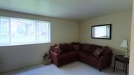 This single-floor space features a living area finished with neutral-toned walls and light carpeting that contributes to a calm, understated atmosphere. The room is anchored by a large sectional sofa in deep burgundy, accented with a mix of patterned and solid throw pillows that add visual interest to the seating arrangement. A large window fitted with vertical blinds allows natural light to filter into the space, brightening the room and offering a view of the greenery outside. A second framed window on the adjacent wall further enhances the sense of openness. The smooth, light-colored ceiling complements the neutral palette throughout. A woven storage basket tucked beside the sofa adds a functional and decorative accent. The overall ambiance of the space is comfortable and relaxed, with a straightforward layout that prioritizes practical living. The carpeted flooring adds warmth underfoot, making the area feel inviting. The room's simplicity allows for flexible furnishing and personalization to suit a variety of lifestyle needs.