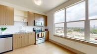 This single-floor unit offers a thoughtfully designed layout that balances functionality and comfort. The kitchen is a standout feature, outfitted with a full suite of stainless steel appliances, including a refrigerator, electric range, over-the-range microwave, and dishwasher. Warm-toned wood cabinetry provides ample storage, while light countertops and a clean backsplash contribute to a modern, streamlined aesthetic. Light wood-look flooring runs throughout much of the unit, lending a bright and airy feel. A large window in the kitchen frames an expansive view of the surrounding cityscape, filling the space with natural light. The bathroom is well-appointed with a wood-finish vanity, granite-style countertop, large mirror, and a full tub-shower combination with tiled surround, maintaining the unit's cohesive modern design. Additional rooms include a carpeted bedroom offering a cozy atmosphere, and an open living area with light wood flooring, crisp white walls, and ceiling vents, providing a blank canvas for personalization. The overall design palette of neutral whites, natural wood tones, and stainless steel creates a clean, contemporary ambiance throughout the space.