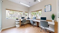 Located at 11615 Firestone Blvd in Norwalk, California, this compact yet efficiently designed business center offers a fully functional shared workspace environment. The room features warm-toned light wood flooring that complements the crisp white and natural wood-accented workstations arranged in an L-shaped configuration along the walls. Multiple desktop computers and a printer are readily available, making the space well-suited for productivity and convenience.

Large windows with wood-trimmed roller shades line two walls, flooding the room with abundant natural light and offering pleasant views of mature trees and the surrounding outdoor greenery. The elevated ceilings, fitted with overhead fluorescent lighting, contribute to an open and airy atmosphere despite the room's cozy footprint.

The neutral, light beige walls are accented by framed artwork, including an abstract landscape painting, adding a polished, welcoming touch to the space. Ergonomic rolling chairs provide comfortable seating at each workstation. A glass partition wall near the entryway hints at an adjacent lounge or common area, seamlessly connecting this business center to the broader facility. Overall, the space strikes a balance between functionality and comfort, offering a professional yet inviting environment for focused work.