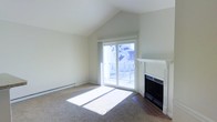This single-floor space offers a welcoming and light-filled interior defined by thoughtful architectural details and a clean, neutral palette. The living area immediately draws the eye upward with its striking vaulted ceiling, which lends a sense of openness and airiness to the room. A fireplace with a crisp white surround serves as a natural focal point, adding warmth and character to the space. Large sliding glass doors flood the interior with generous natural light, casting bright, sun-drenched patterns across the carpeted floor throughout the day. These doors open directly onto a private balcony, offering a seamless connection between the indoor living area and the outdoors. The baseboard heating system runs along the exterior wall, ensuring comfort throughout the seasons. The neutral tones of the walls and flooring create a versatile and inviting atmosphere, well-suited to a variety of interior styles. Overall, the space combines practical features with architectural charm, resulting in a comfortable and visually appealing single-level layout that feels both spacious and intimate.