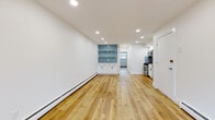 This single-floor unit showcases a thoughtfully renovated interior with a clean, modern aesthetic defined by crisp white walls, recessed lighting, and warm-toned hardwood floors that run continuously throughout the space. The open layout creates a generous living area with an airy feel, complemented by baseboard heating along the perimeter walls. A set of glass patio doors at one end floods the space with natural light and provides outdoor access.

The kitchen is efficiently designed with white shaker cabinetry, a herringbone-patterned teal tile backsplash, white countertops, and a suite of stainless steel appliances including a gas range, over-the-range microwave, and refrigerator. Open shelving with the same teal tile backdrop extends into the living area, serving as a decorative and functional built-in display area with lower cabinet storage below.

A hallway connects the main living space to additional rooms, including a bathroom finished with slate-gray floor tiles, white cabinetry, matte black hardware with brass accents, and blue tile detailing in the shower. The overall design balances contemporary finishes with practical functionality throughout.