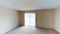 This single-floor space presents a clean, functional layout centered around a spacious living area. The room is finished with wall-to-wall carpeting in a neutral beige tone, complementing the warm, light-colored walls that create a cohesive and understated aesthetic throughout. A textured popcorn ceiling adds a classic touch overhead, while standard white baseboards frame the perimeter of the room cleanly.

A prominent sliding glass door serves as the focal point of the main living area, fitted with vertical blinds that allow for adjustable light control and privacy. The door opens to an outdoor balcony, offering a view of a green outdoor setting and allowing natural light to filter generously into the interior. The balcony railing is visible through the glass, suggesting a modest but practical outdoor extension of the living space.

Practical details include a floor-level HVAC vent positioned near the sliding door for efficient climate control, as well as a wall-mounted vent near the ceiling, ensuring even air distribution. Standard electrical outlets are conveniently placed along the walls. Overall, the space offers a simple, well-lit environment suitable for comfortable everyday living.