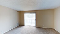 This single-floor space presents a clean, functional layout centered around a spacious living area. The room is finished with wall-to-wall carpeting in a neutral beige tone, complementing the warm, light-colored walls that create a cohesive and understated aesthetic throughout. A textured popcorn ceiling adds a classic touch overhead, while standard white baseboards frame the perimeter of the room cleanly.

A prominent sliding glass door serves as the focal point of the main living area, fitted with vertical blinds that allow for adjustable light control and privacy. The door opens to an outdoor balcony, offering a view of a green outdoor setting and allowing natural light to filter generously into the interior. The balcony railing is visible through the glass, suggesting a modest but practical outdoor extension of the living space.

Practical details include a floor-level HVAC vent positioned near the sliding door for efficient climate control, as well as a wall-mounted vent near the ceiling, ensuring even air distribution. Standard electrical outlets are conveniently placed along the walls. Overall, the space offers a simple, well-lit environment suitable for comfortable everyday living.