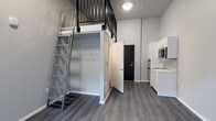 This studio with one bathroom and a loft is a smartly designed living space that makes efficient use of its footprint. Dark hardwood-style flooring runs throughout, providing a warm contrast to the crisp grey and white walls that keep the interior feeling bright and clean. The highlight of the main living area is the elevated loft, accessible via a sturdy metal ladder with a sleek industrial aesthetic. The loft level is framed by a black metal railing, offering a dedicated sleeping or lounging area that keeps the floor below open and functional. Directly beneath the loft is a built-in open closet with wire shelving, providing organized storage without occupying additional floor space. The compact kitchen is positioned along one wall and features white cabinetry, a stainless steel sink, and a built-in microwave, delivering essential cooking and prep functionality in a streamlined layout. A black-framed entry door adds a bold design accent that complements the industrial touches seen in the ladder and loft railing. The bathroom is conveniently situated off the main living area. The overall design of this space prioritizes clean lines, practical storage solutions, and a modern aesthetic, making it a well-organized and stylish living environment.