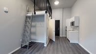 This studio with one bathroom and a loft is a smartly designed living space that makes efficient use of its footprint. Dark hardwood-style flooring runs throughout, providing a warm contrast to the crisp grey and white walls that keep the interior feeling bright and clean. The highlight of the main living area is the elevated loft, accessible via a sturdy metal ladder with a sleek industrial aesthetic. The loft level is framed by a black metal railing, offering a dedicated sleeping or lounging area that keeps the floor below open and functional. Directly beneath the loft is a built-in open closet with wire shelving, providing organized storage without occupying additional floor space. The compact kitchen is positioned along one wall and features white cabinetry, a stainless steel sink, and a built-in microwave, delivering essential cooking and prep functionality in a streamlined layout. A black-framed entry door adds a bold design accent that complements the industrial touches seen in the ladder and loft railing. The bathroom is conveniently situated off the main living area. The overall design of this space prioritizes clean lines, practical storage solutions, and a modern aesthetic, making it a well-organized and stylish living environment.