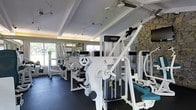 This single-floor fitness center is designed to provide a fully functional workout environment with a wide array of professional-grade equipment. The space is lined with white strength training machines, including cable systems, leg press units, and various resistance machines, all arranged to maximize accessibility and flow throughout the room. The dark rubber flooring provides a durable, slip-resistant surface suited for high-traffic exercise use. Natural light pours in through large windows along the perimeter walls, creating a bright and energizing atmosphere while offering views of the surrounding greenery outside. A striking natural stone accent wall adds a distinctive architectural character to the space, contrasting with the clean white tones of the equipment and ceiling. The ceiling features elegant coffered detailing with recessed lighting and track lighting, ensuring the space is well-lit at all times. Ceiling fans contribute to comfortable air circulation during workouts. Mirrored walls on one side enhance the sense of depth and openness while serving a functional purpose for form monitoring. Overall, the space balances a welcoming aesthetic with practical, professional fitness facility design.