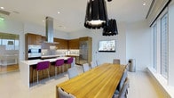 This single-floor space showcases a thoughtfully designed open-plan layout that integrates the kitchen and dining areas into one cohesive, contemporary environment. The kitchen is outfitted with warm wood cabinetry, a stainless steel refrigerator, and a sleek range hood that anchors the cooking zone. A white countertop island with vibrant purple bar stools serves as both a prep area and casual seating space, adding a striking pop of color against the neutral palette.

The dining area features a large, solid wood rectangular table surrounded by an eclectic mix of upholstered and woven chairs, comfortably accommodating a generous number of guests. A wall-mounted flat-screen television positioned nearby adds a functional media element to the entertaining space.

Overhead, a cluster of dramatic black drum pendant lights draws the eye upward, emphasizing the soaring ceiling height and lending bold visual contrast to the bright white walls and light-toned tile flooring. Recessed lighting supplements the statement pendants, ensuring the space is well-illuminated throughout. The overall ambiance is modern, clean, and well-appointed, balancing functionality with a strong design aesthetic.