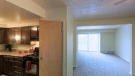 This single-floor residential space offers a practical and comfortable layout designed for everyday living. The open living area is finished with plush wall-to-wall carpeting that adds warmth underfoot, while a large sliding glass door dressed with vertical blinds floods the room with natural light and provides a bright, airy atmosphere. A suspended tile ceiling runs throughout, lending a clean and consistent look across the space. Adjacent to the living area, the kitchen is partially separated by a dividing wall, offering a semi-open feel while maintaining a distinct cooking zone. The kitchen is equipped with dark, rich cabinetry that contrasts with the lighter walls, and features a gas range, providing a functional setup for home cooking. The overall ambiance of the space is understated and practical, with neutral tones throughout that offer a versatile backdrop for a variety of interior styles. The thoughtful separation between the kitchen and living areas allows for comfortable daily use while maintaining an open and connected feel throughout the single-level floor plan.