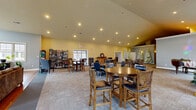 Located at 48 Bavarian Way in Altamont, NY, this expansive single-floor community clubhouse offers a generous and versatile layout designed for socializing and recreation. The space comprises three distinct living areas, a hallway, and a welcoming entrance, all spread across a substantial footprint that encourages both intimate gatherings and larger community events.

The main gathering areas feature warm carpeted flooring, vaulted and cathedral ceilings, and recessed lighting that create an open, inviting ambiance. Multiple high-top tables and chairs are arranged throughout, complemented by a well-stocked bookshelf lending a cozy, lounge-like atmosphere. Large windows throughout allow ample natural light to fill the rooms.

The kitchen area is bright and functional, finished with cool blue walls, a large granite-topped center island with an integrated sink and dishwasher, stainless steel appliances, and crisp white cabinetry — ideal for catering and community events.

A dedicated media room features plush recliner seating, a large wall-mounted flat-screen television, and a built-in sound system, providing an intimate entertainment experience.

The game room rounds out the amenity offerings with hardwood-style flooring, a billiards table, a ping pong table, a ceiling fan, and large windows overlooking the surrounding community — a bright and energetic space for recreation.