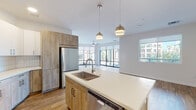 This single-floor space showcases a contemporary design with a well-considered open-concept layout connecting the kitchen, dining, and living areas. The kitchen is a standout feature, equipped with a large center island topped with white quartz countertops that includes an undermount sink and dishwasher. Cabinetry combines white upper cabinets with warm wood-toned lower cabinets, complemented by a white tile backsplash and a full suite of stainless steel appliances. Pendant lighting in brushed gold adds a refined finishing touch above the island.

The living area flows seamlessly from the kitchen and is bathed in natural light through expansive floor-to-ceiling windows and sliding glass doors, offering views of palm trees and the surrounding urban landscape. Wood-look flooring runs throughout the main living spaces, while carpeted bedrooms provide a softer feel. Each bedroom features a ceiling fan and windows framing outdoor views.

The bathrooms are finished with wood-toned vanities, white quartz countertops, large mirrors, and wood-look flooring. One bathroom includes a tub-shower combination, while another features a walk-in shower. The overall aesthetic is clean, modern, and cohesive throughout.