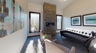 This thoughtfully designed single-floor space serves as a welcoming and functional common area, blending modern aesthetics with warm, inviting details. The entry lobby immediately impresses with soaring double-height ceilings and expansive floor-to-ceiling glass doors and windows that flood the space with natural light, offering clear views to the surrounding exterior. Large-format light gray tile flooring anchors the entrance, creating a clean and contemporary foundation. A comfortable seating area near the entry features tufted upholstered chairs arranged around a small accent table, with a framed pass-through window offering a glimpse into an adjacent bar or lounge space with counter-height seating. Beyond the entryway, a carpeted lounge area provides a cozy retreat centered around a dramatic stacked-stone fireplace that rises to the ceiling, flanked by dark-framed windows that allow natural light to filter in from multiple angles. The warm tones of the stone contrast elegantly with the neutral walls and soft furnishings, including a tufted sofa and accent chairs. A glass-enclosed conference room with a sliding dark-framed door offers a private, professional workspace visible from the corridor, furnished with a large table and ergonomic seating. Throughout the space, curated artwork and carefully selected décor contribute to a polished, upscale ambiance.