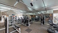 Located at 2555 Summer Dr in Dublin, Ohio, this well-appointed fitness center offers a comprehensive workout environment on a single floor. The space is defined by its striking vaulted ceiling with a grid-paneled design, which adds architectural interest while contributing to an open, airy atmosphere. Full-length mirrored walls line the perimeter, visually expanding the space and allowing users to monitor their form throughout their workouts. The flooring features durable, textured carpeting with designated mat zones that help organize the workout areas and provide traction underfoot. The facility is outfitted with an extensive selection of strength training equipment, including cable machines, weight benches, and a range of selectorized resistance machines suited for targeting various muscle groups. Cardio equipment, including ellipticals and steppers, is positioned along the walls, complementing the strength training offerings. Overhead, recessed and grid-mounted lighting ensures the entire floor is well-illuminated. The layout is thoughtfully arranged to allow for comfortable circulation between stations, making the space functional for multiple users simultaneously. Overall, the fitness center presents a clean, professional environment designed to support a full range of exercise routines.