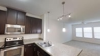 This single-floor space offers a thoughtfully designed open-concept layout that seamlessly connects the kitchen and living areas, creating a bright and functional environment. The kitchen is a standout feature, showcasing rich dark espresso cabinetry that provides generous storage and a striking visual contrast against the light-toned laminate countertops. A double-basin stainless steel sink is positioned along the peninsula, making it a practical hub for meal prep and everyday tasks. Above the peninsula, two sleek cylindrical pendant lights hang at varying heights, adding a contemporary decorative touch. A track lighting fixture further illuminates the adjoining living space, ensuring consistent, even lighting throughout. Stainless steel appliances, including a built-in microwave, complement the kitchen's modern aesthetic. The adjacent living area transitions smoothly with neutral-toned flooring and crisp white walls, contributing to an airy, clean ambiance. A window allows natural light to filter into the living space, enhancing the open feel of the unit. The textured ceiling and clean lines throughout lend the space a polished, move-in-ready character well-suited for modern living.