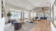 Located at 12202 Huffmeister Road in Cypress, Texas, this single-floor community space offers an open-concept layout that seamlessly integrates a living room, dining area, and kitchen. The interior is defined by dramatic vaulted ceilings that amplify the sense of openness throughout, while an abundance of large windows floods the space with natural light and frames views of an outdoor pool area.

The living room features multiple distinct seating arrangements, including plush sofas, accent chairs, and geometric wood coffee tables set atop neutral area rugs. Built-in shelving niches with teal-green backing add a decorative focal point alongside a wall-mounted display screen. Wood-look plank flooring transitions seamlessly across the main areas, contributing warmth and continuity.

The kitchen is appointed with dark navy cabinetry, quartz countertops, stainless steel appliances, and a full-height whitewashed brick accent wall that adds textural contrast. Pendant lantern lights hang above the elongated island, reinforcing the modern-yet-inviting aesthetic. The adjacent dining area offers additional seating with a refined, intimate atmosphere. Together, these spaces create a cohesive, well-appointed environment suited for gathering and everyday use.