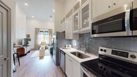 This thoughtfully designed single-floor residence offers a cohesive open-concept layout that seamlessly connects the kitchen and living area. The kitchen is a standout feature, showcasing white shaker-style cabinetry that extends toward impressively tall ceilings, complemented by glass-front upper cabinets, a blue-gray subway tile backsplash, white quartz countertops, and a full suite of stainless steel appliances. Warm wood-look flooring runs throughout, unifying the space with a consistent, modern aesthetic. The living area is bright and welcoming, bathed in natural light from large windows dressed with elegant linen drapes. Soaring ceiling heights lend the space an airy, open feel. The bedroom continues the clean, serene design language with large windows framing outdoor views, white walls, and soft natural light. The bathroom features a white tile surround, light tile flooring, a gray vanity with an undermount sink, and a well-lit mirror, offering a crisp and functional retreat. Near the kitchen entry, a louvered closet door and a well-appointed entryway provide convenient storage solutions. Throughout, the space balances modern finishes with warm, livable touches, creating an environment that is both stylish and comfortable.