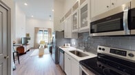 This thoughtfully designed single-floor residence offers a cohesive open-concept layout that seamlessly connects the kitchen and living area. The kitchen is a standout feature, showcasing white shaker-style cabinetry that extends toward impressively tall ceilings, complemented by glass-front upper cabinets, a blue-gray subway tile backsplash, white quartz countertops, and a full suite of stainless steel appliances. Warm wood-look flooring runs throughout, unifying the space with a consistent, modern aesthetic. The living area is bright and welcoming, bathed in natural light from large windows dressed with elegant linen drapes. Soaring ceiling heights lend the space an airy, open feel. The bedroom continues the clean, serene design language with large windows framing outdoor views, white walls, and soft natural light. The bathroom features a white tile surround, light tile flooring, a gray vanity with an undermount sink, and a well-lit mirror, offering a crisp and functional retreat. Near the kitchen entry, a louvered closet door and a well-appointed entryway provide convenient storage solutions. Throughout, the space balances modern finishes with warm, livable touches, creating an environment that is both stylish and comfortable.