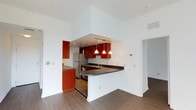 This single-floor residence offers a thoughtfully designed layout with an emphasis on openness and functionality. The kitchen serves as a central highlight, featuring rich cherry-toned wood cabinetry that provides ample storage, complemented by dark countertops and stainless steel appliances, including a full-sized refrigerator. A curved breakfast bar extends from the kitchen, creating a natural division between the cooking and living areas while offering casual seating and additional workspace.

Three pendant lights hang above the kitchen area, casting warm, focused illumination that enhances the space's inviting ambiance. The kitchen is partially recessed beneath a soffit, adding architectural depth and definition to the open floor plan.

The living area benefits from soaring ceilings that amplify the sense of space, while crisp white walls provide a clean, neutral backdrop throughout. Durable hardwood-style flooring runs consistently across the space, lending warmth and visual continuity. A hallway leads to additional rooms, suggesting a well-organized and practical floor plan. The combination of elevated ceilings, open sightlines, and warm wood tones creates a balanced and welcoming atmosphere throughout this well-appointed home.